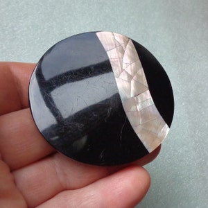 May include: A round, black brooch with a diagonal inlay of iridescent, cracked mother-of-pearl. The brooch is held in a hand, showcasing its polished surface and unique design. The jewelry piece is a striking accessory.