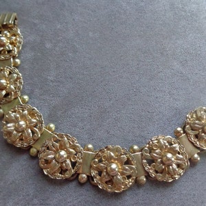 May include: Gold-toned bracelet featuring a floral design. The bracelet is composed of multiple circular floral elements connected by rectangular links and small spherical beads. The clasp is a simple hook and bar closure. The jewelry is set against a gray background.