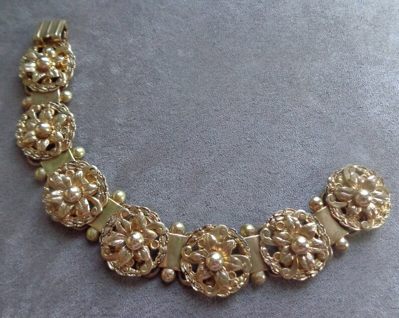 May include: Gold-toned bracelet featuring a floral design. The bracelet is composed of multiple circular floral elements connected by rectangular links and small spherical beads. The clasp is a simple hook and bar closure. The jewelry is set against a gray background.