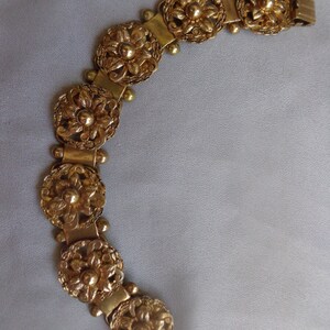 May include: A gold-toned bracelet featuring a series of ornate, circular floral designs. Each circular element is connected by rectangular links and small, spherical accents. The bracelet has a flexible, articulated design, suggesting it can be worn comfortably.