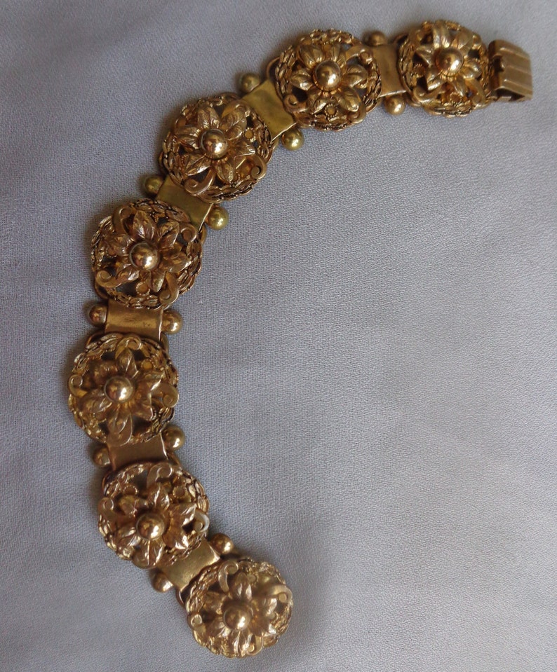 May include: A gold-toned bracelet featuring a series of ornate, circular floral designs. Each circular element is connected by rectangular links and small, spherical accents. The bracelet has a flexible, articulated design, suggesting it can be worn comfortably.