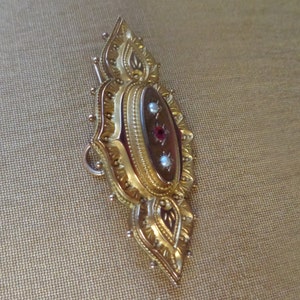 May include: A gold-toned brooch with an oval-shaped cameo set in a detailed, ornate frame. The cameo features a red stone surrounded by two smaller white stones.