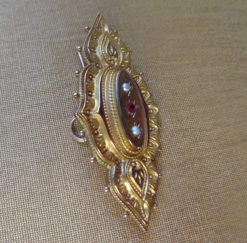 May include: A gold-toned brooch with an oval-shaped cameo set in a detailed, ornate frame. The cameo features a red stone surrounded by two smaller white stones.