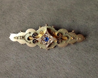 May include: A gold brooch featuring a blue gemstone surrounded by smaller white gemstones. The brooch has a detailed design with intricate swirls and beading.