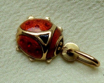 Puede incluir: Un dije de oro con forma de mariquita con alas de esmalte rojo que presentan pequeños puntos oscuros y un acento triangular negro cerca de la cabeza. El dije incluye una pequeña argolla circular de oro en la parte superior para colgar.