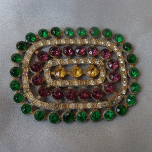 Puede incluir: Un broche vintage con un diseño de múltiples capas con piedras de imitación verdes, moradas y amarillas engastadas en un marco de metal plateado.