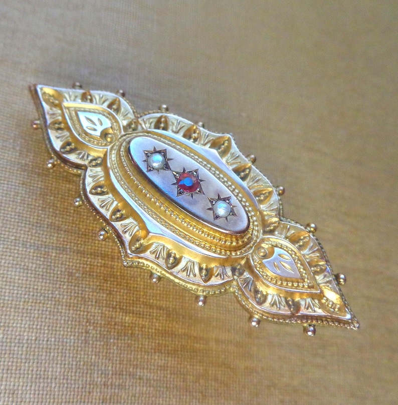 May include: A gold brooch with a white cameo set in a gold filigree frame. The cameo features three red and white gemstones.