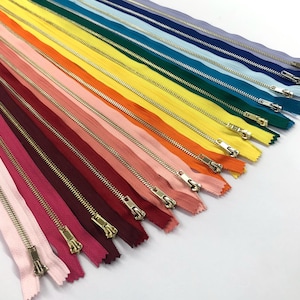 May include: A collection of 20 different colored zipper closures with gold metal pulls. The zippers are arranged in a rainbow pattern, with the darkest colors at the bottom and the lightest colors at the top.