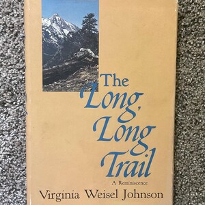 May include: A hardcover book titled The Long, Long Trail: A Reminiscence by Virginia Weisel Johnson. The tan cover features a rectangular photograph of a snow-capped mountain peak and a pine tree. The title is printed in large blue serif font.