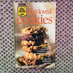 May include: A cookbook titled "Nestle Toll House Best-Loved Cookies" featuring a stack of chocolate chip cookies. The cover includes the text "70 most requested cookies, bars and brownies" and "15 tips for foolproof cookies".