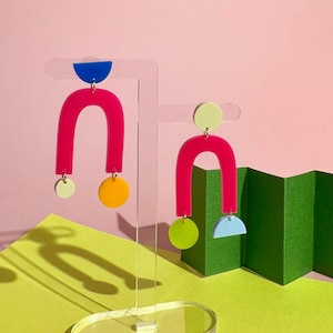 Peut inclure: Paire de boucles d'oreilles abstraites sur un présentoir transparent. Les boucles d'oreilles présentent un design rose vif en forme de U avec des formes géométriques colorées attachées en bleu, jaune et vert. Le fond est un mélange de rose, de vert et de jaune.