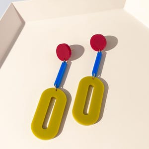 May include: A pair of drop earrings featuring a red circular stud, a blue rectangular connector, and a large olive green oval pendant with a central cutout. The components are linked together with small gold-toned metal jump rings.