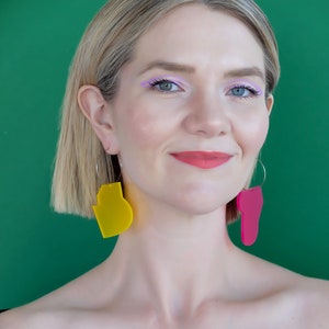 May include: A woman with blonde hair and blue eyes wearing large, abstract, asymmetrical earrings. One earring is yellow, the other is pink. The background is a solid green. The woman is wearing a black top and has pink lipstick and purple eyeliner.
