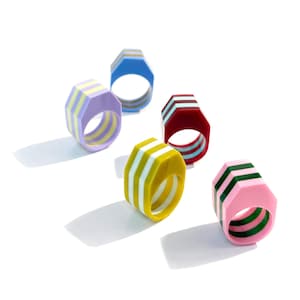 May include: A collection of colorful, geometric rings. Each ring features a unique combination of colors, including pink, yellow, blue, and burgundy, with striped detailing. The rings are set against a white background, highlighting their vibrant design.