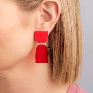 May include: Vibrant red, geometric earrings. Each earring has a half-circle shape attached to a larger, rounded rectangle. The earrings hang from a small gold-coloured wire. A striking, contemporary accessory.