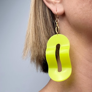 May include: Bright neon yellow statement earrings with a gold-coloured chain and hook. The earrings have a unique, abstract shape with a cutout in the centre. A bold, eye-catching accessory.