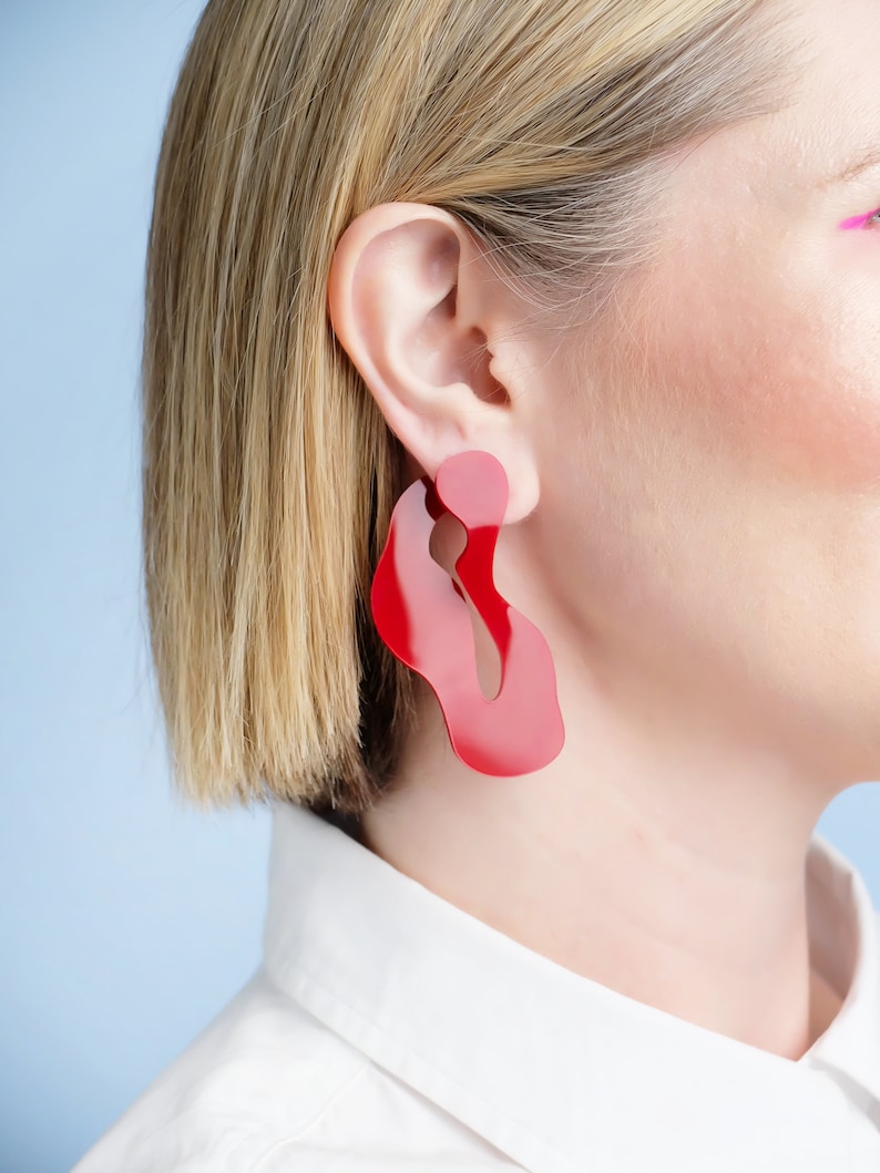 Pode incluir: Um par de brincos vermelhos abstratos grandes. Os brincos t&ecirc;m um acabamento brilhante e uma forma &uacute;nica e fluida. O fundo &eacute; azul claro e a pessoa est&aacute; usando uma camisa branca com gola.