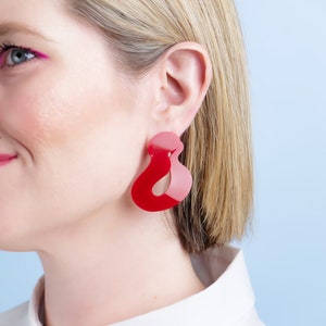 Pode incluir: Um close-up de uma pessoa usando um par de brincos grandes, abstratos, vermelhos e rosa. Os brincos t&ecirc;m um formato &uacute;nico e fluido e s&atilde;o feitos de um material brilhante. A pessoa est&aacute; usando uma camisa branca com gola.