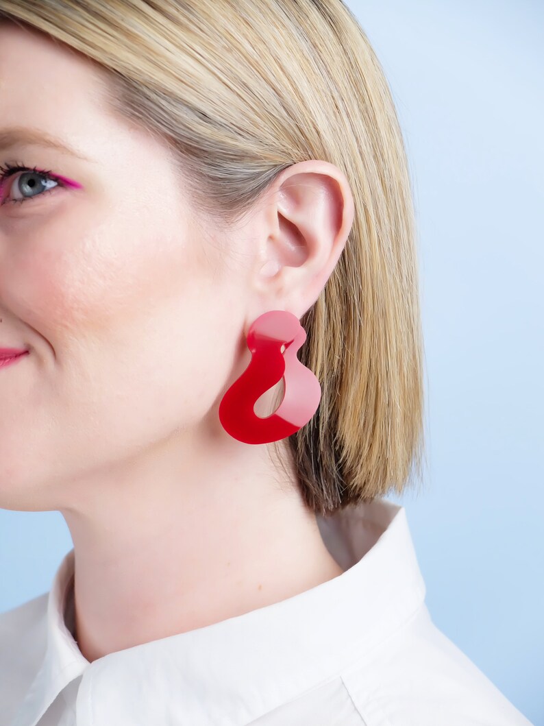 Pode incluir: Um close-up de uma pessoa usando um par de brincos grandes, abstratos, vermelhos e rosa. Os brincos t&ecirc;m um formato &uacute;nico e fluido e s&atilde;o feitos de um material brilhante. A pessoa est&aacute; usando uma camisa branca com gola.