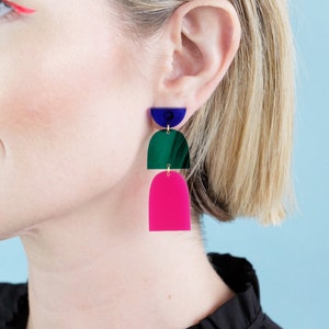 May include: Geometric dangle earrings in blue, green, and hot pink. The earrings feature a layered design with a half-circle, a curved arch, and a rectangular arch. The earrings are suspended from a gold-coloured hook.