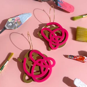 May include: Two pairs of abstract earrings with gold hoops. The earrings have a layered design of olive green and hot pink shapes. Paintbrushes and palette knives with colourful paint are scattered on a pink background.