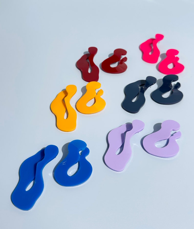 Pode incluir: Uma cole&ccedil;&atilde;o de brincos de acr&iacute;lico coloridos em v&aacute;rias tonalidades, incluindo vermelho, rosa, amarelo, azul, roxo e azul marinho. Os brincos t&ecirc;m um design abstrato e ondulado. Os brincos s&atilde;o exibidos em uma superf&iacute;cie branca.