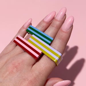 May include: Three colorful striped rings in red, blue, and yellow are displayed. The rings have a rectangular shape with parallel stripes. The background is a solid pink color.