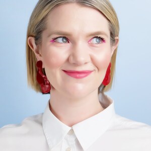 Pode incluir: Uma pessoa usando brincos vermelhos grandes e abstratos. A pessoa est&aacute; usando uma camisa branca com gola e tem delineador e batom rosa. O fundo &eacute; azul claro.