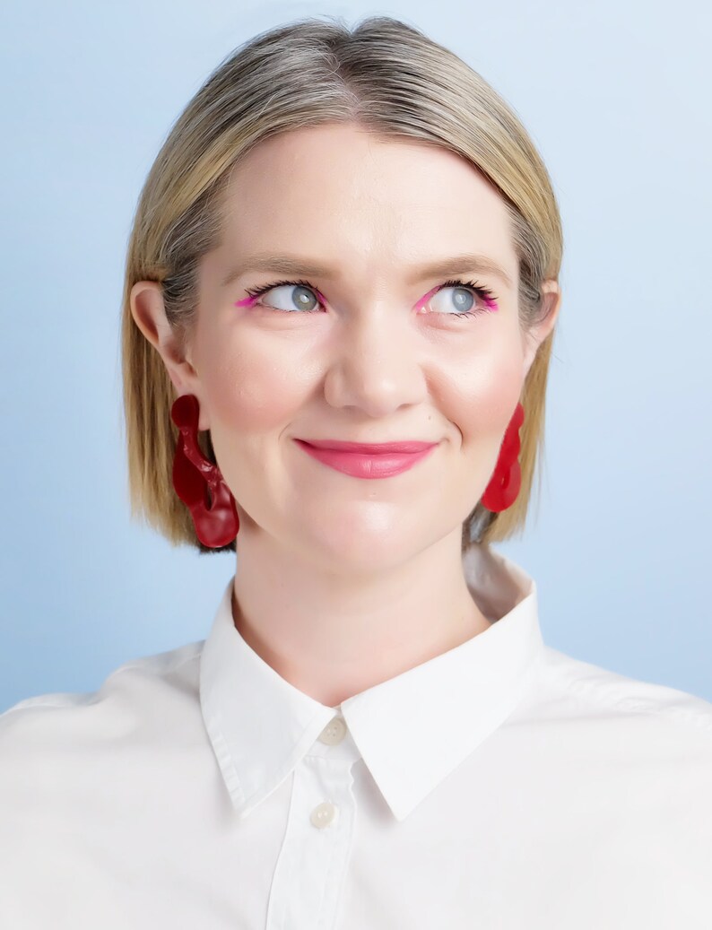 Pode incluir: Uma pessoa usando brincos vermelhos grandes e abstratos. A pessoa est&aacute; usando uma camisa branca com gola e tem delineador e batom rosa. O fundo &eacute; azul claro.