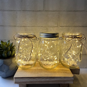 May include: Three clear glass mason jars with lids, each containing a string of white fairy lights. The jars are sitting on a wooden tray with a rustic finish.
