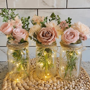 May include: Three clear glass mason jars with pink and peach-colored flowers and greenery. Each jar is wrapped with twine and contains fairy lights. The jars are arranged on a woven, circular placemat, creating a rustic floral arrangement.