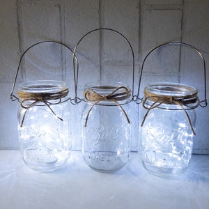 May include: Three clear glass mason jars with metal handles and twine bows. Each jar contains a string of white fairy lights.