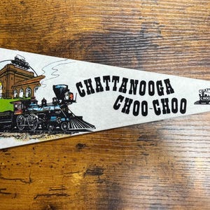May include: A white pennant with a red border and yellow tabs. The pennant features a vintage illustration of a train, a building, and the text "Chattanooga Choo Choo". The pennant also includes the text "Chattanooga Tennessee".