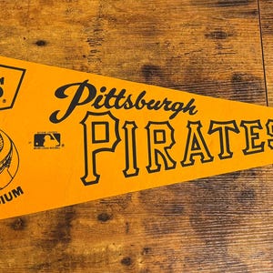 May include: A pennant in the shape of a triangle, featuring the words "Pittsburgh Pirates" in black lettering on a yellow background. The pennant also includes the Denny's logo and an illustration of Three Rivers Stadium.