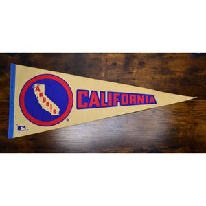 May include: A tan triangular felt pennant featuring a blue circle with a red border. Inside the circle is a white map of California with the word Angels written vertically in red. The word CALIFORNIA is printed in large blue letters to the right.