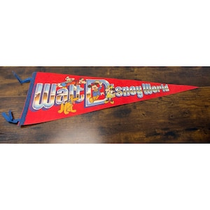 May include: A red triangular felt pennant featuring the text Walt Disney World in large blue and white gradient lettering. The design includes colorful illustrations of Mickey Mouse, Minnie Mouse, Donald Duck, Goofy, and Pluto.
