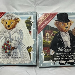 May include: Two teddy bear paper dolls, one dressed as a bride and the other as a groom. The bride doll is wearing a white dress with lace trim and a veil. The groom doll is wearing a black tuxedo with a white shirt and bow tie. Both dolls have a Steiff/Strong museum label.