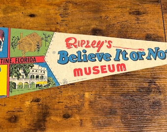 May include: Vintage pennant featuring the text "Ripley's Believe It or Not! Museum." The pennant has illustrations of St. Augustine, Florida, including a donkey, a log cabin, and a building. The pennant is white with red, blue, yellow, and green accents.