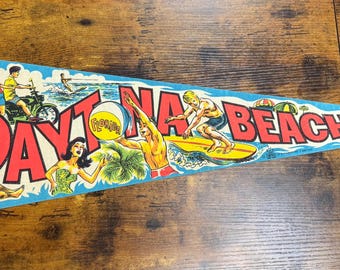 May include: A vintage pennant flag with a blue background and the words "Daytona Beach" in red and white. The pennant features illustrations of people surfing, riding a motorcycle, and playing with a beach ball. The pennant is trimmed in green and yellow.