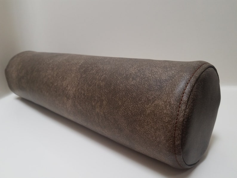 May include: A brown cylindrical pillow with a textured fabric cover. The pillow has a rounded end and is sewn with a dark brown thread.