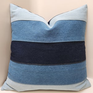 May include: A blue and white striped denim pillow cover. The pillow cover is made of different shades of blue denim sewn together in horizontal stripes.