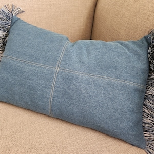 May include: A rectangular denim pillow featuring a medium blue wash with a cross-shaped seam pattern stitched in gold thread. The left and right edges of the pillow are finished with long, multi-tonal blue and cream-colored fabric fringe.