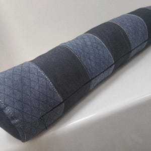 May include: A cylindrical bolster pillow with a denim patchwork design. The pillow features alternating sections of solid and patterned denim in shades of blue and gray, with white stitching. The pillow is approximately 24 inches long.