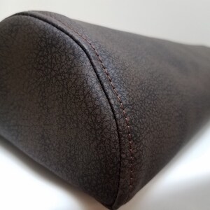 May include: A brown faux leather lumbar support pillow with a stitched seam around the edge.