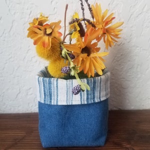 May include: A small, square denim fabric container with a striped fabric lining, holding artificial yellow and orange flowers and berries. The container is a decorative item, suitable for holding small plants or other items.