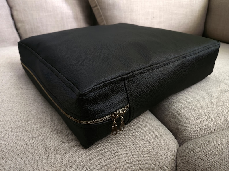 K&ouml;nnte beinhalten: Schwarzes Lederkissen mit Rei&szlig;verschluss. Das Kissen liegt auf einem hellgrauen Sofa.
