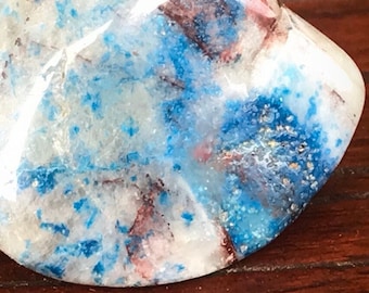 May include: A polished, tumbled gemstone featuring a mottled pattern of vibrant blue, creamy white, and translucent gray sections with thin, dark brown mineral inclusions throughout the smooth, rounded, and reflective surface of the stone.