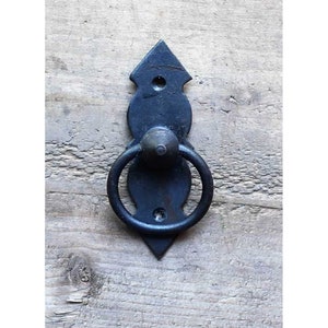 Könnte beinhalten: Schwarzer Metallring-Zuggriff mit dekorativem Design. Der Griff ist an einer Holzoberfläche befestigt.