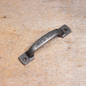 Peut inclure: Une poignée en fonte noire avec les mots "CHEESE BOARD" en relief. La poignée est conçue pour être fixée à une planche à fromage en bois.