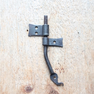 May include: Black metal door latch with a hook-shaped handle and two plates with holes. The latch is attached to a wooden surface.